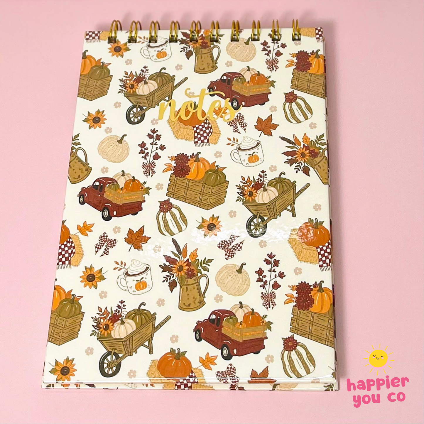 An autumn-themed spiral-bound notepad with a cream background and a pattern of pumpkins, fall leaves, harvest carts, and coffee cups, displayed on a solid pink background.