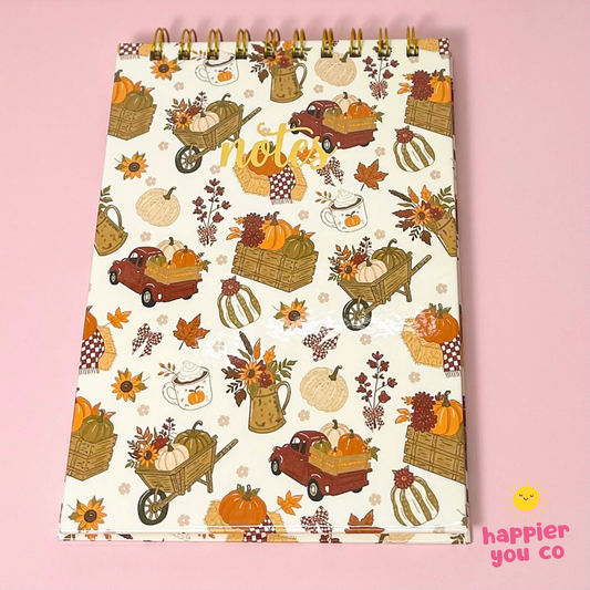 An autumn-themed spiral-bound notepad with a cream background and a pattern of pumpkins, fall leaves, harvest carts, and coffee cups, displayed on a solid pink background.