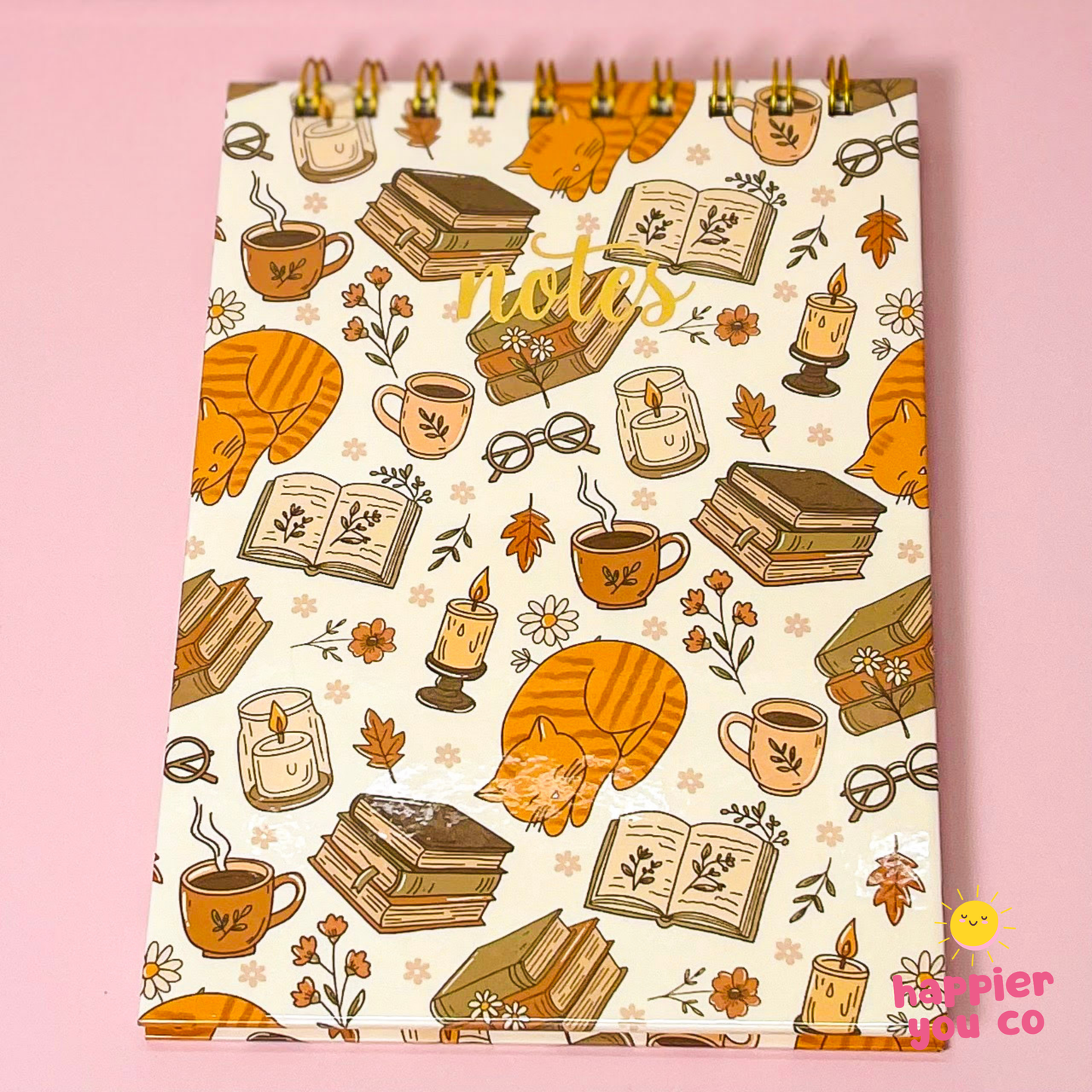 A spiral-bound notepad with a cozy autumn and owl-themed pattern, featuring orange tabby owls, books, candles, and coffee cups on a cream background, set against a solid pink backdrop.