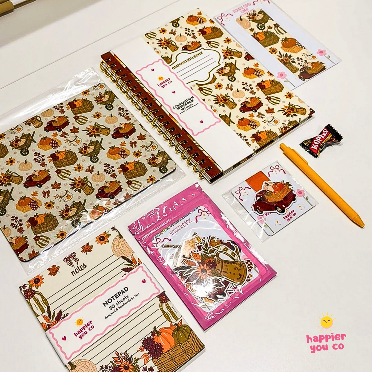 A flat-lay collection of autumn-themed stationery items on a white surface, including patterned notebooks, notepad pads, sticker sheets, a yellow pen, and a small candy wrapper. The stationery features fall motifs like pumpkins, leaves, and baskets in orange, brown, and beige tones.