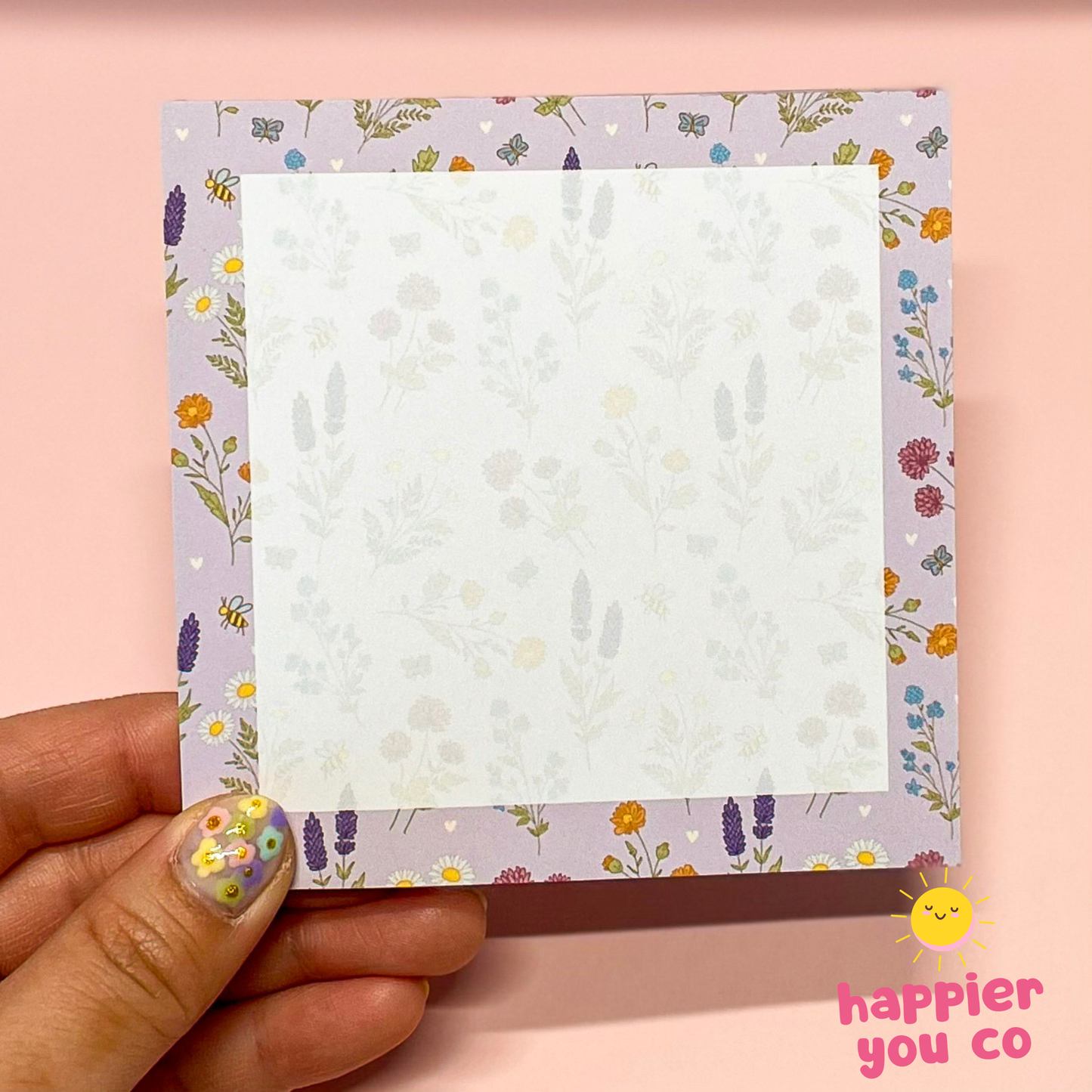 Hand holding a notepad with floral border on a pink background, featuring the brand 'happier you co'.