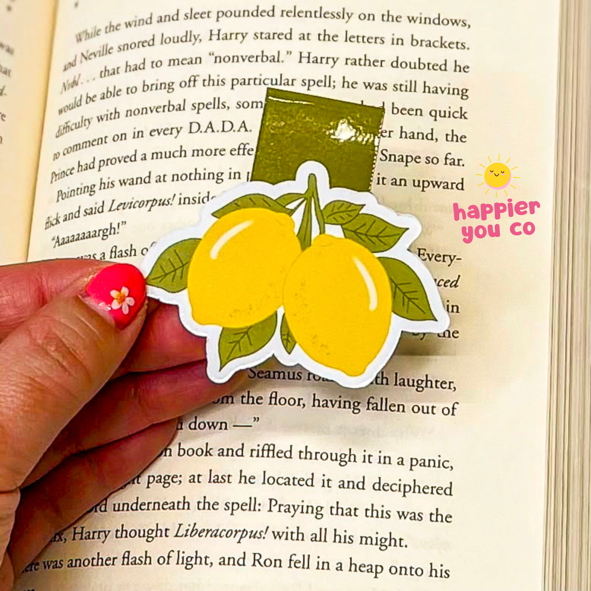 Hand holding a sticker of lemons on an open book with 'happier you co' branding.