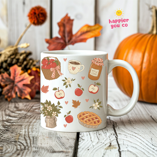 A white ceramic mug decorated with autumn-themed illustrations, including apples, a pie, a cup of coffee, and a basket of red apples, set on a rustic wooden table with a pumpkin and fall leaves in the background. The brand logo 'happier you co' is visible in the top right corner.