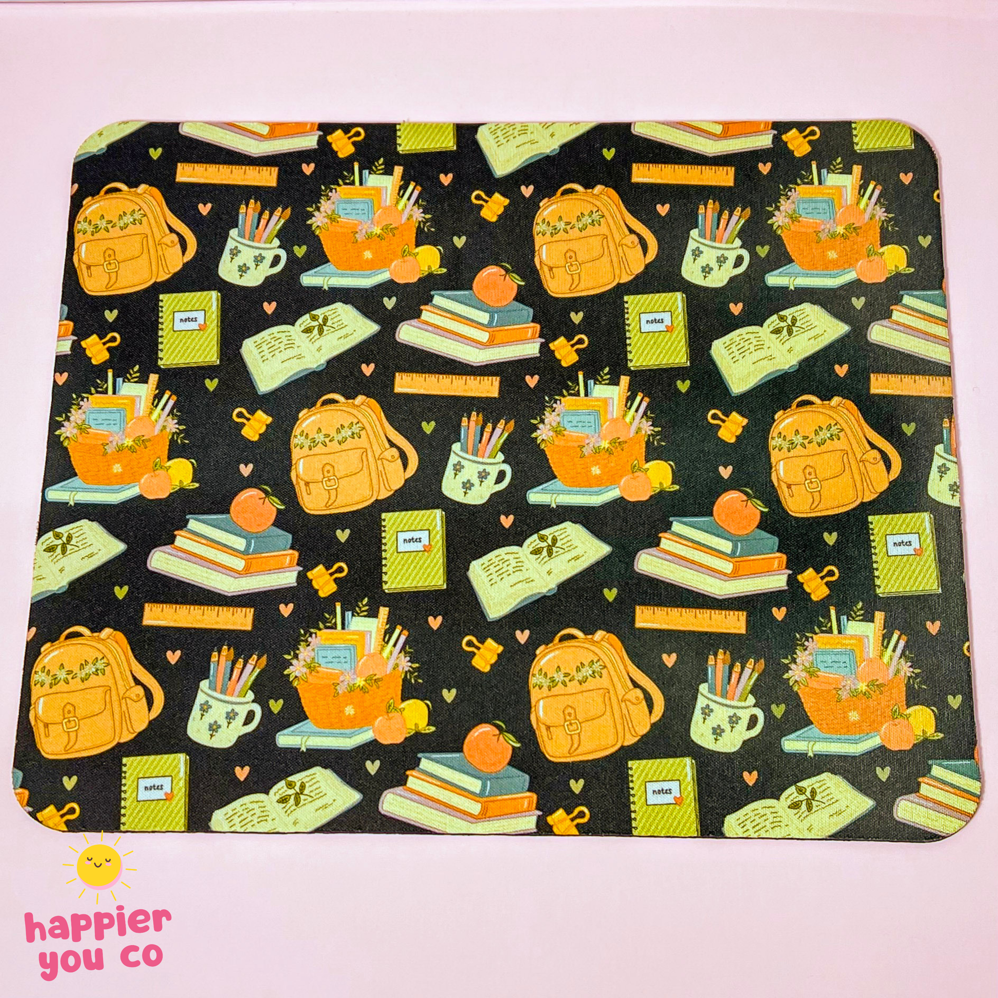 A rectangular desk mat with rounded corners featuring a black background and a colorful school-themed pattern of backpacks, books, notebooks, pencils, and mugs. The desk mat is photographed flat on a light pink background, with a 'happier you co' logo in the bottom-left corner.