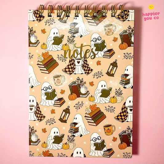 A spiral-bound notebook with a warm beige cover featuring a repeating autumn-themed pattern of reading ghosts, books, pumpkins, mugs, and lanterns, with the word 'notes' in script across the center. The notebook is photographed against a solid pink background, with a small 'happier you co' logo in the top right corner.