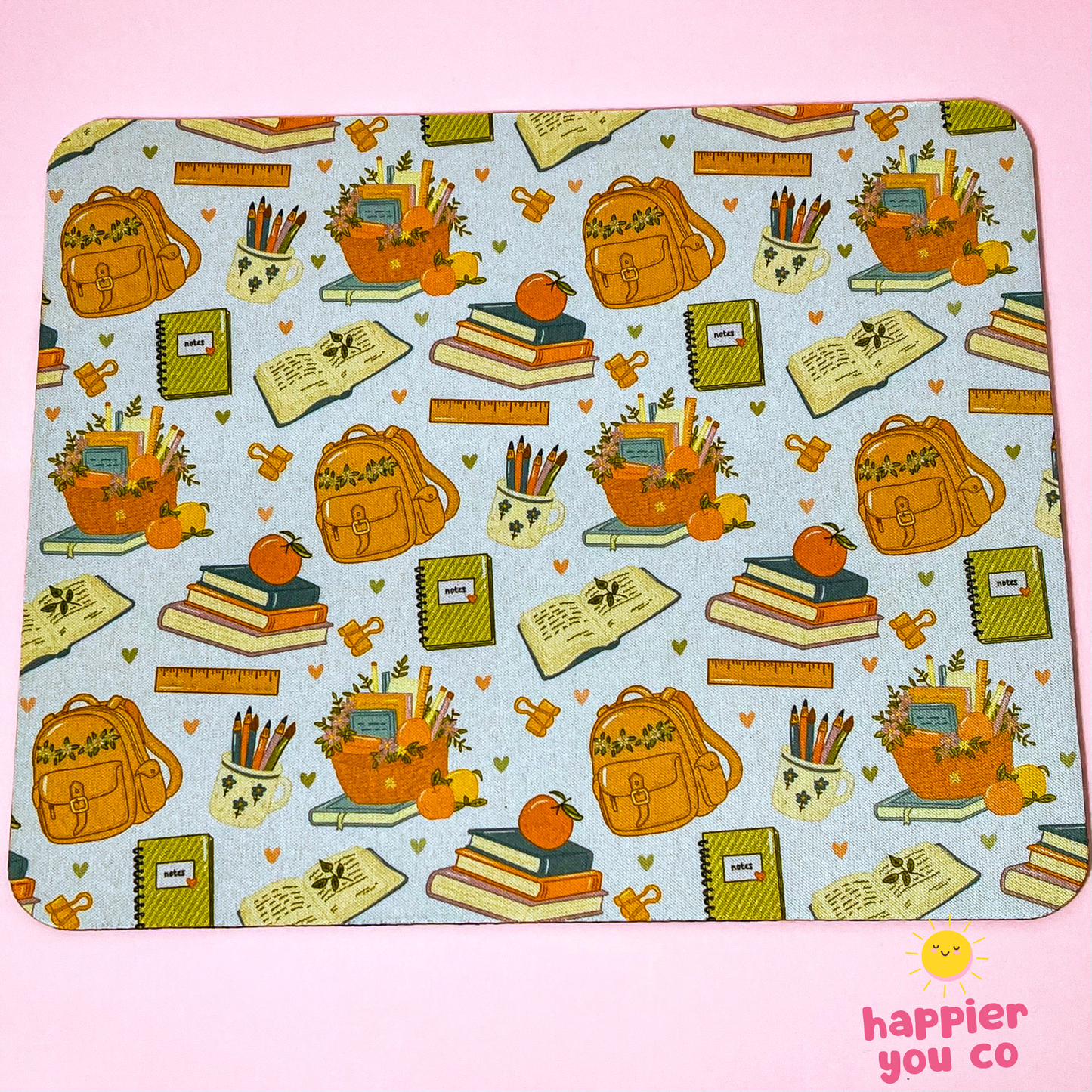 A rectangular mousepad with rounded corners featuring a repeating autumn school-themed pattern on a light blue background. The pattern includes orange backpacks, stacks of books with pumpkins on top, notebooks, pencils, and small hearts.