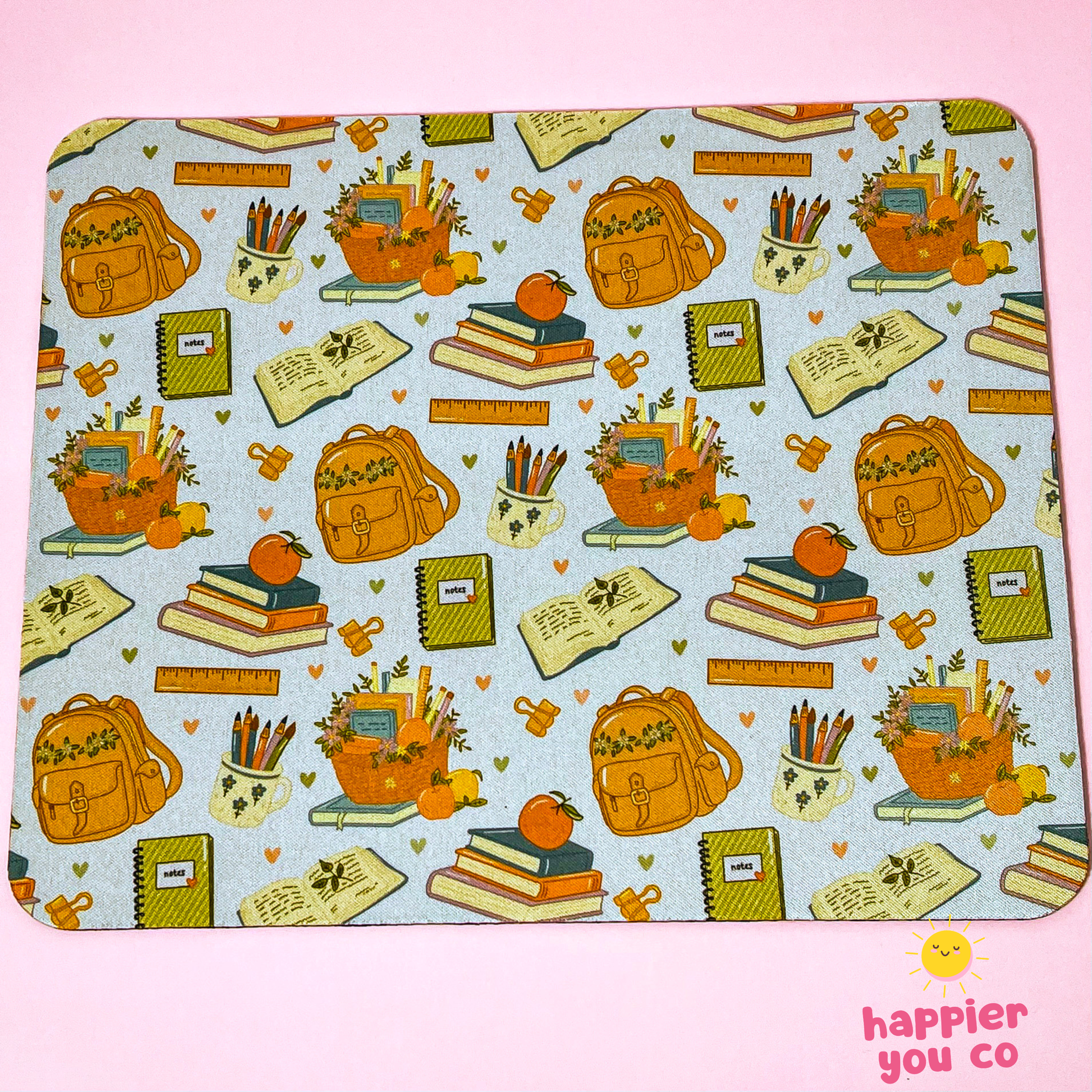 A rectangular mousepad with rounded corners featuring a repeating autumn school-themed pattern on a light blue background. The pattern includes orange backpacks, stacks of books with pumpkins on top, notebooks, pencils, and small hearts.