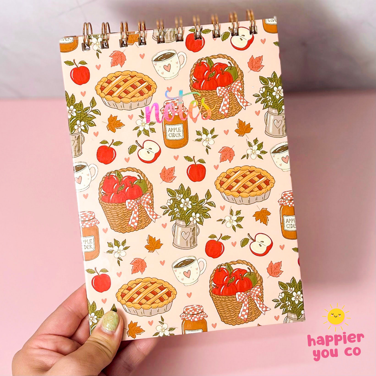A hand holds a spiral-bound notebook with a warm autumn pattern featuring red apples, pies, coffee cups, and baskets, set against a soft pink background.