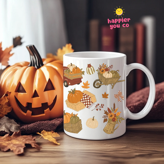 A white ceramic mug with a colorful autumn harvest print of pumpkins, gourds, leaves, and baskets, styled on a wooden table next to a carved jack-o'-lantern pumpkin with a 'happier you co' logo in the upper right.