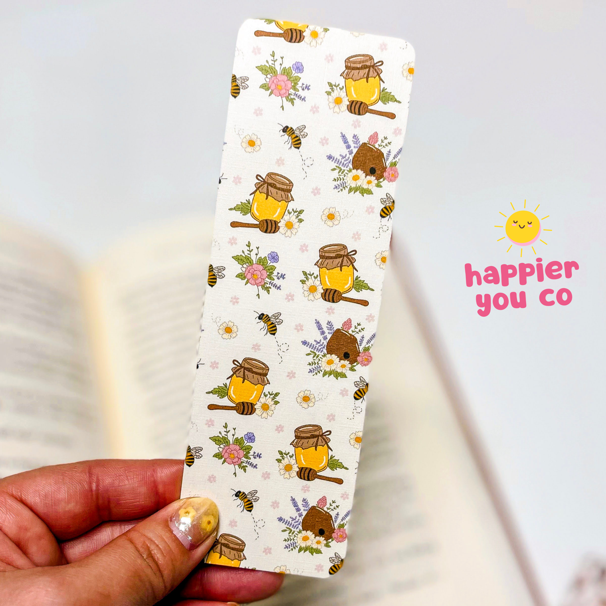 Bookmark with bee and honeycomb design held by a hand, 'happier you co' logo visible.