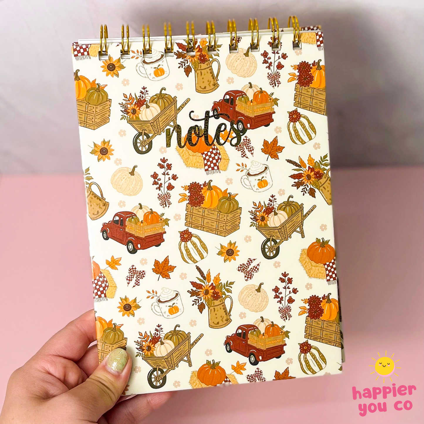 A hand holding a spiral-bound notebook with an autumn-themed cover featuring pumpkins, autumn leaves, flowers, and the word 'notes' in script. The background is a soft pink surface with a light gray wall above.