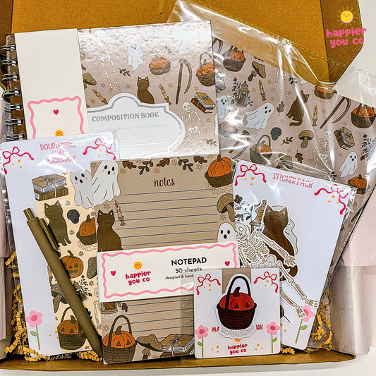 A Halloween-themed stationery gift set arranged in a box, including a composition book, notepad, sticker sheet, memo pad, and pen, all decorated with pumpkins, ghosts, skeletons, and bows. The brand 'happier you co' is visible on several items.