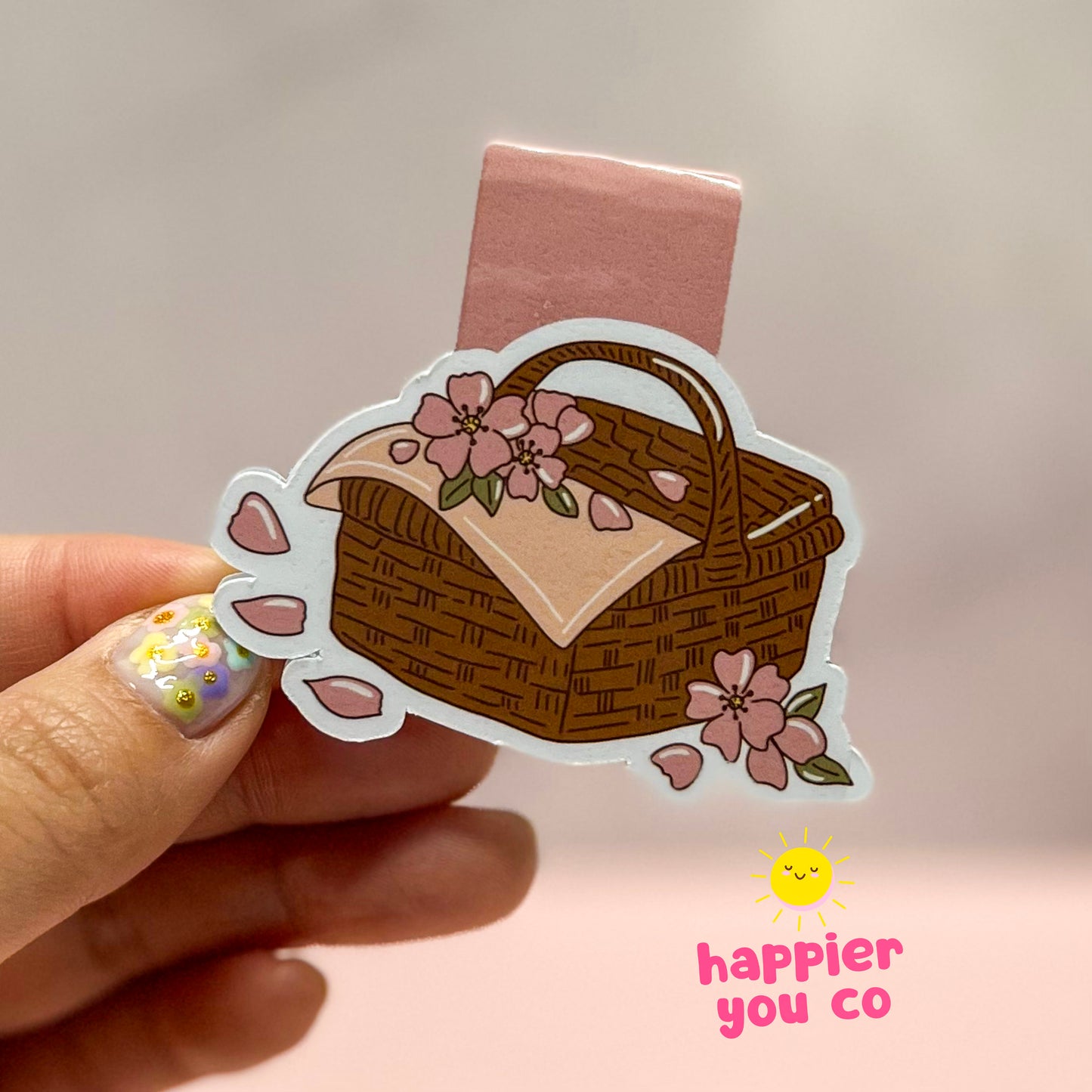 Magnetic bookmark of a picnic basket with flowers held by a hand, brand 'happier you co' visible.