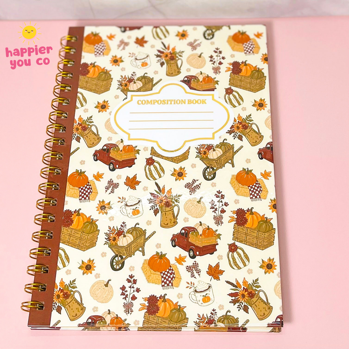An autumn-themed composition notebook with a cream cover decorated with pumpkins, baskets, leaves, and vintage trucks, bound with a brown paper ribbon and gold spiral. The notebook is placed on a light pink background, with a 'happier you co' logo in the top-left corner.