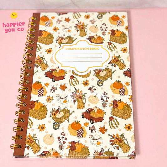 An autumn-themed composition notebook with a cream cover decorated with pumpkins, baskets, leaves, and vintage trucks, bound with a brown paper ribbon and gold spiral. The notebook is placed on a light pink background, with a 'happier you co' logo in the top-left corner.
