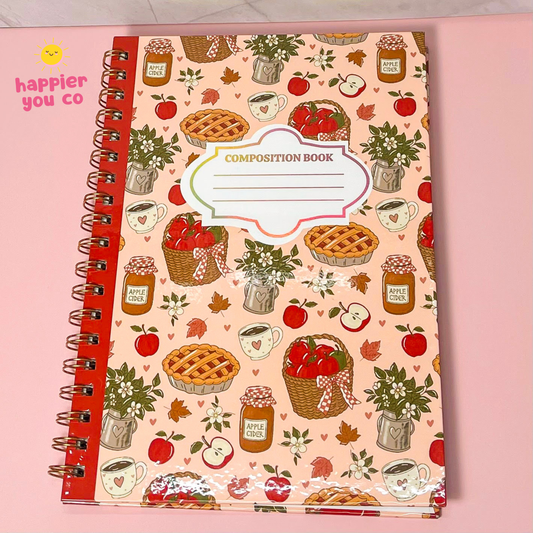 A spiral-bound composition notebook with a pink cover decorated with an autumn picnic pattern of pies, apple cider, jars, and plants, placed on a pink background.