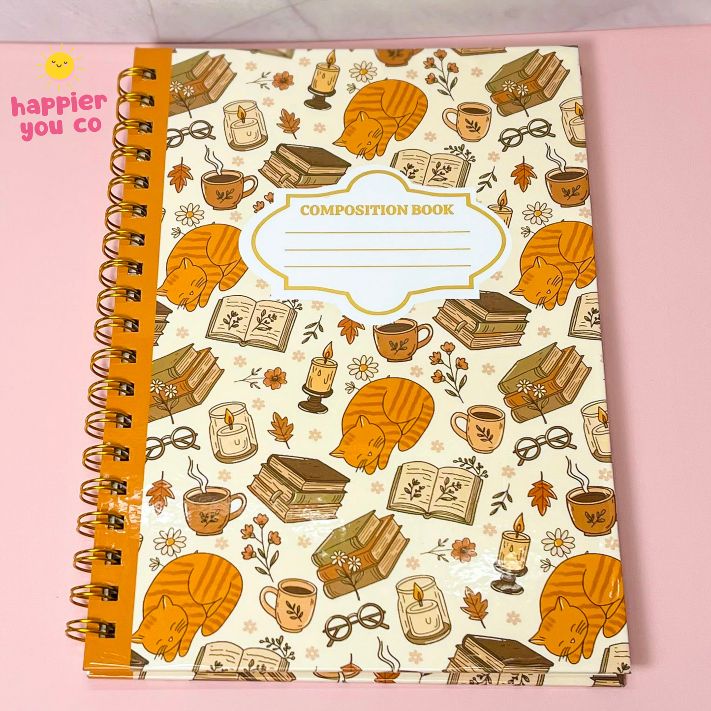 A spiral-bound composition notebook with a cream cover decorated with orange sleeping cats, books, candles, coffee cups, and autumn leaves. The notebook is photographed on a pink background, with a 'happier you co' logo in the top-left corner.
