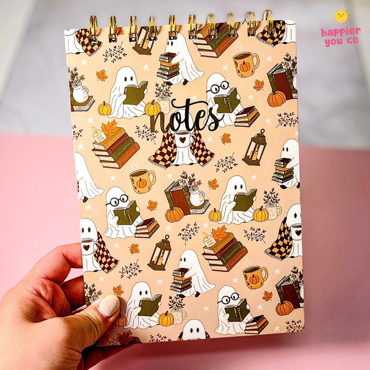 A hand holds a spiral-bound notebook with a Halloween-themed cover featuring ghosts, books, pumpkins, and autumn motifs on a beige background. The brand name 'happier you co' is visible in the top right corner.