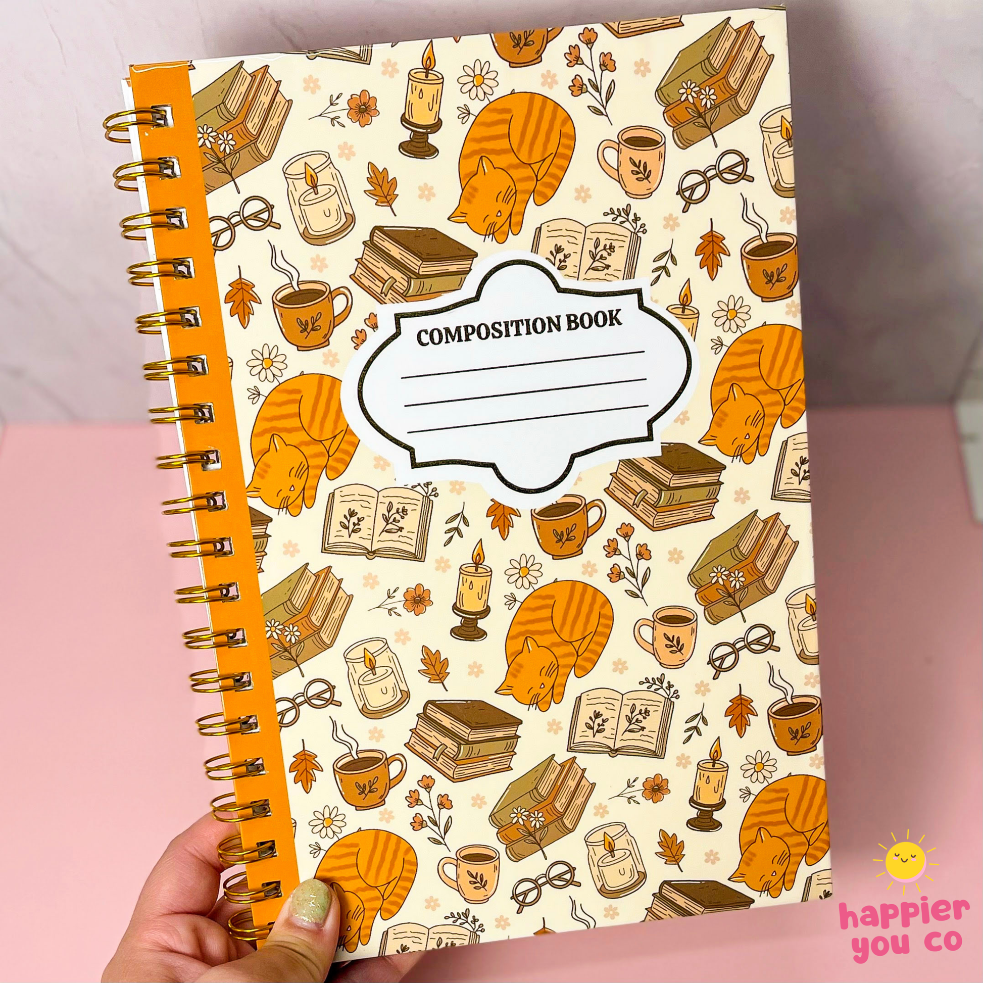 A hand holding a spiral-bound composition notebook with a cream cover decorated with an autumn-themed pattern of orange cats, books, candles, and coffee cups. The notebook has a bright orange spine and gold spiral binding.