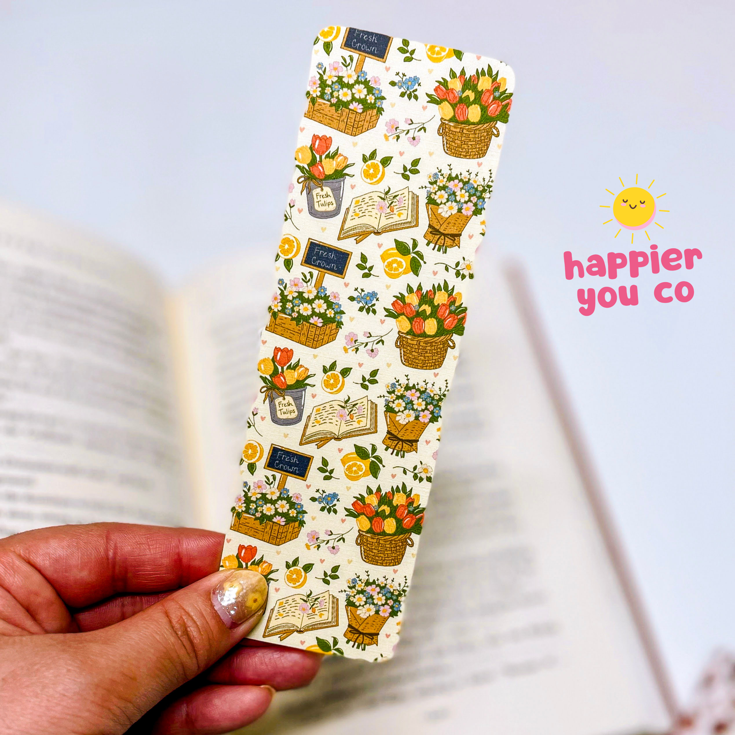 Decorative bookmark with floral and book illustrations held by a hand, with 'happier you co' branding.