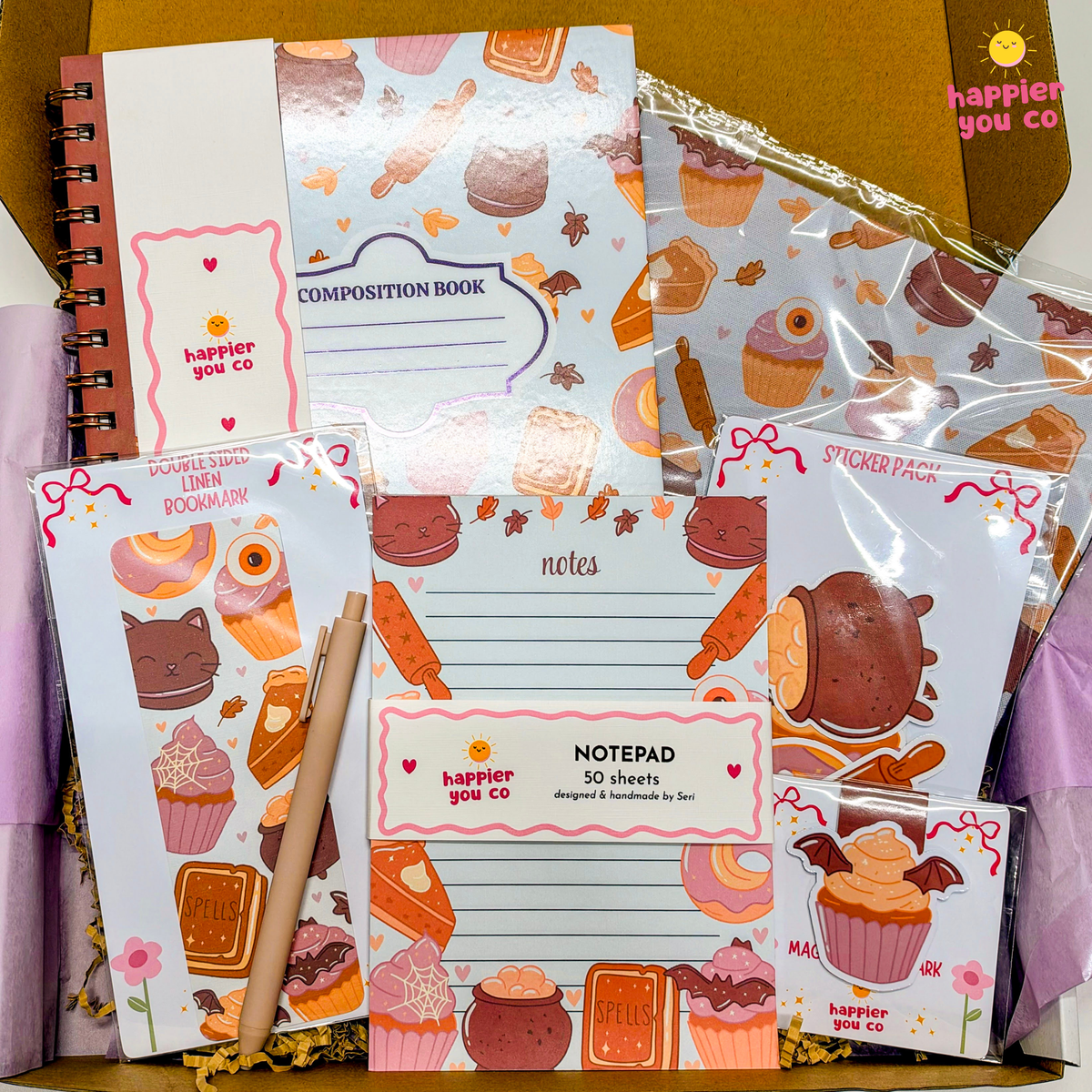 A curated flat-lay of bakery-themed stationery items inside a gift box, including a composition book, notepad, sticker sheets, and a pen, all decorated with cute cupcake and pastry illustrations. The brand name 'happier you co' is visible on several items.