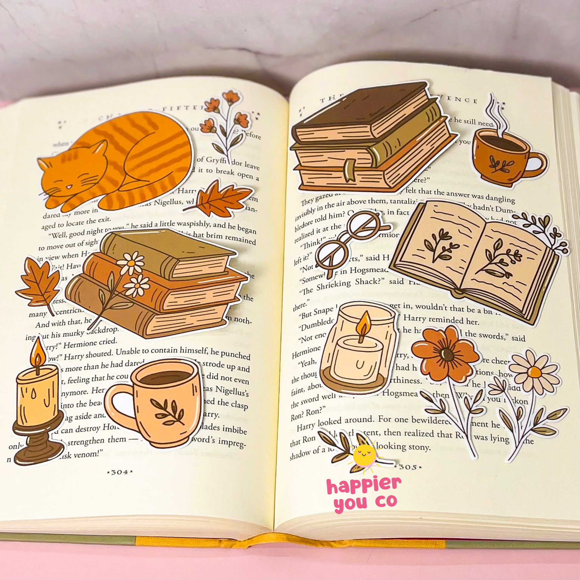 An open decorative book with illustrated autumn and cozy themes, including an orange cat, stacked books, a mug, candles, flowers, and small drawings layered over printed text. The phrase 'happier you co' appears in pink at the bottom of the right page.