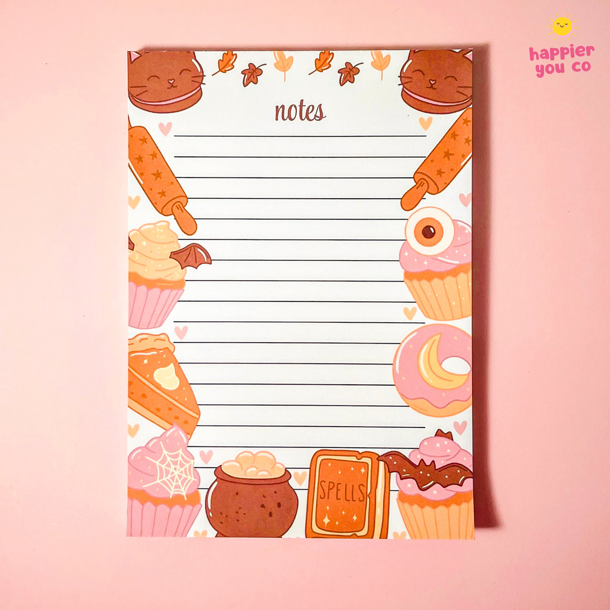 A cute Halloween-themed lined notepad with illustrations of cupcakes, a slice of cake, a cauldron, a spellbook, and a rolling pin, displayed on a solid pink background.