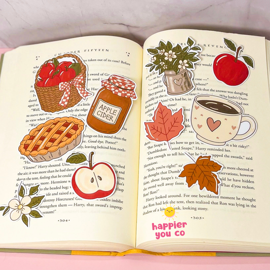 A decorative book page sticker set featuring autumn and apple-themed illustrations, shown applied to the pages of an open book. The stickers include a basket of red apples, a jar of apple cider, a apple pie, a red apple, white flowers, a heart-patterned mug with a red apple, red maple leaves, and a brown autumn leaf.
