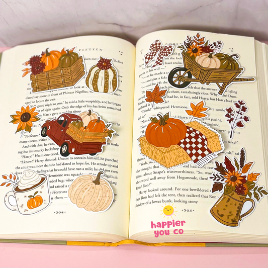 An open book spread decorated with autumn-themed die-cut stickers, including pumpkins, fall leaves, a wheelbarrow, a wagon, a mug, and a pumpkin patch, placed on a pink surface.