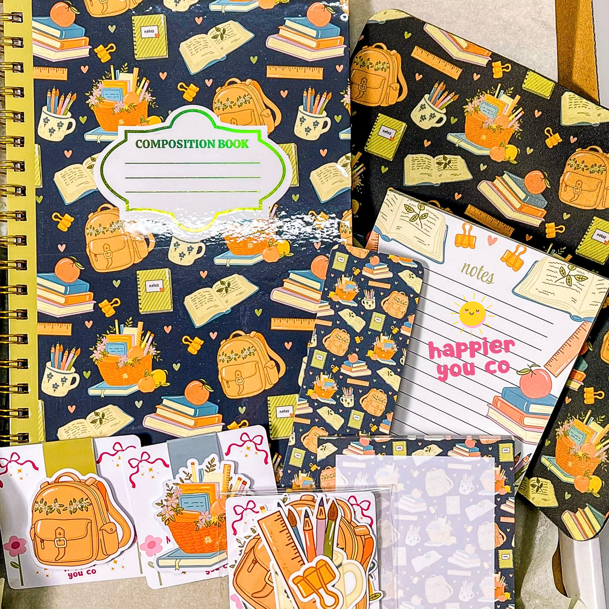 A flat lay of school-themed stationery items, including a composition notebook, a notepad, stickers, and cards, all featuring a navy blue background with a colorful pattern of backpacks, books, pencils, and coffee cups.