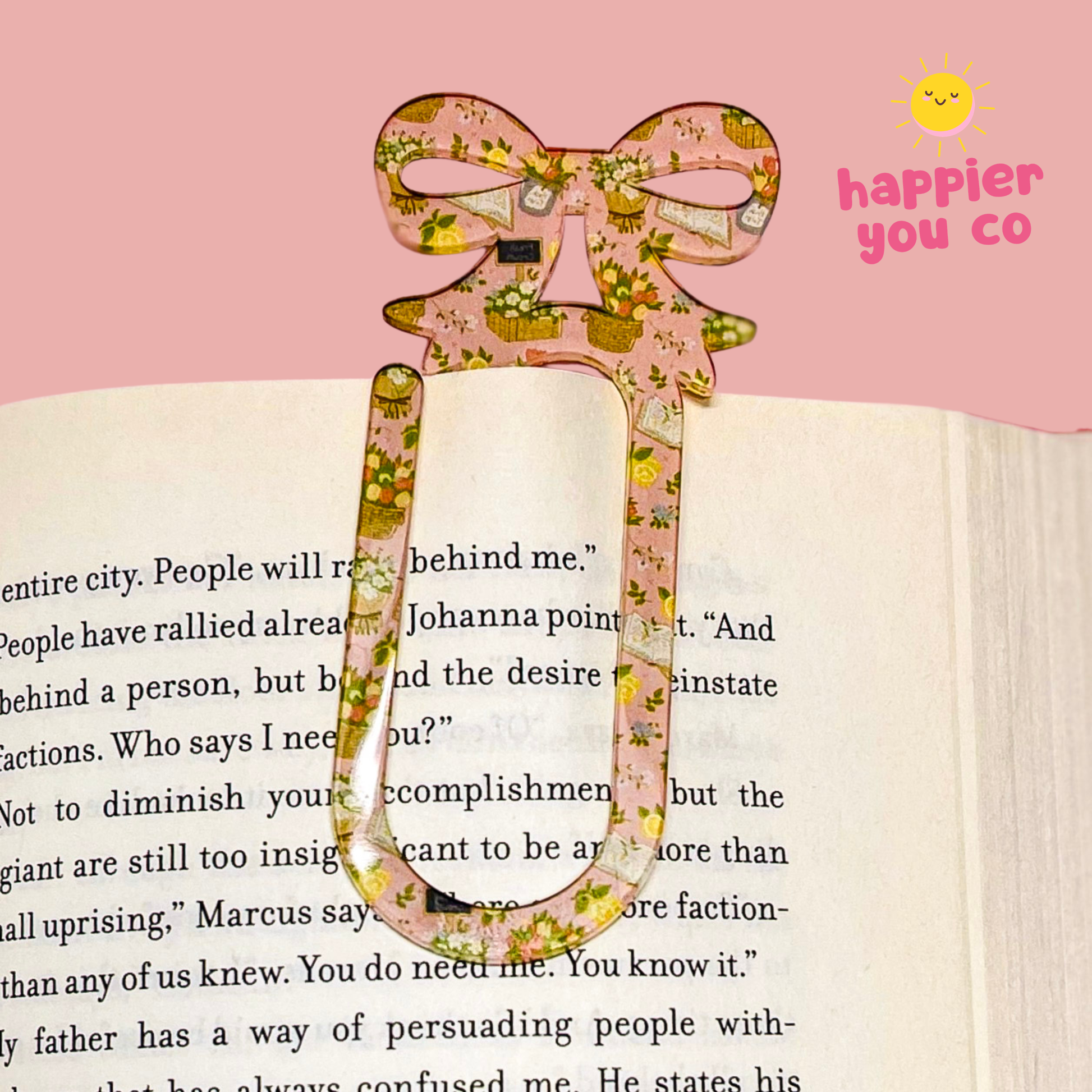 Floral bookmark on an open book with a pink background and 'happier you co' text.
