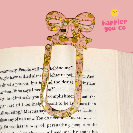 Floral bookmark on an open book with a pink background and 'happier you co' text.