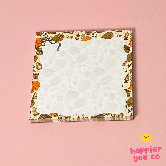 A square notepad with a patterned border featuring autumn-themed illustrations like pumpkins, leaves, and mugs, set against a solid pink background. A 'happier you co' logo is visible in the bottom right corner.