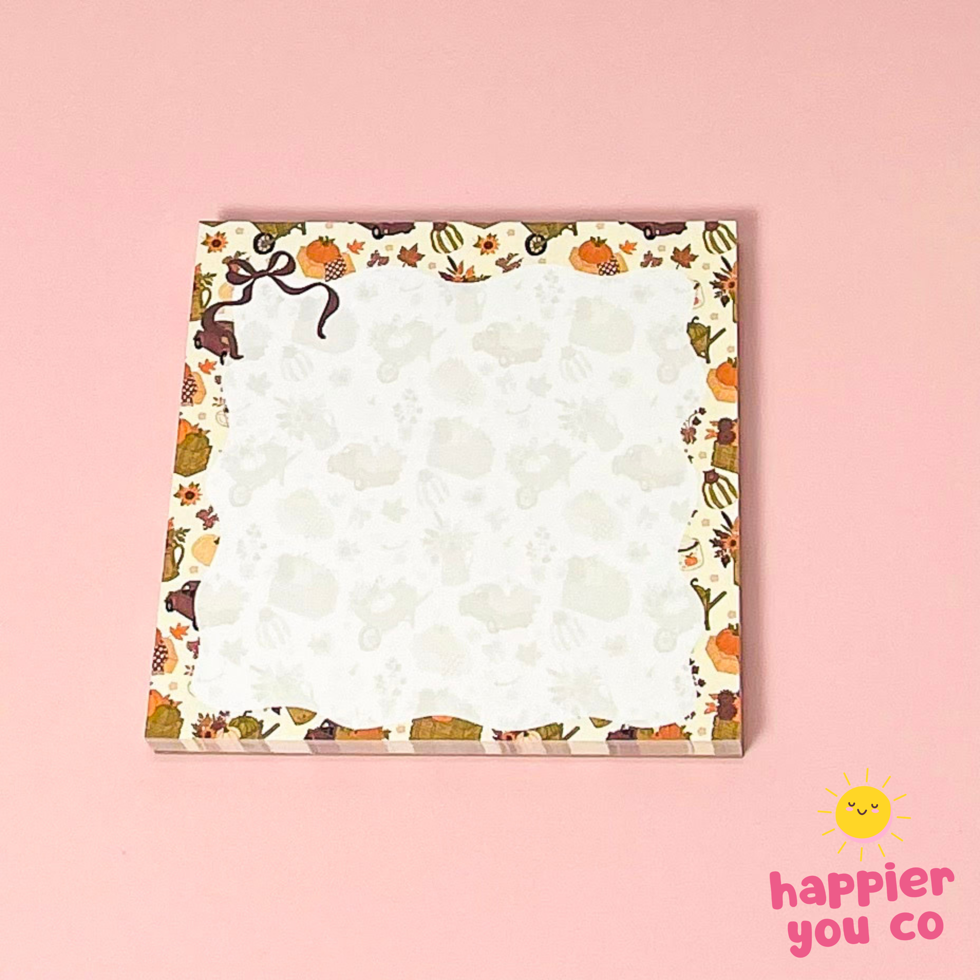 A square notepad with a cute autumn-themed border featuring pumpkins, leaves, and animals on a cream background, set against a solid pink backdrop. The 'happier you co' logo is visible in the bottom right corner.