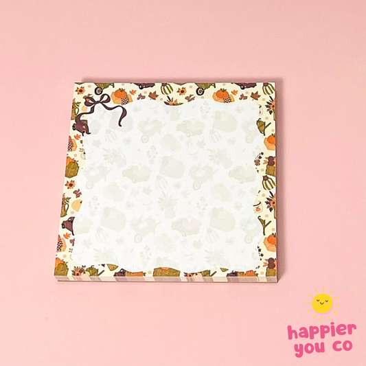 A square notepad with a cute autumn-themed border featuring pumpkins, leaves, and animals on a cream background, set against a solid pink backdrop. The 'happier you co' logo is visible in the bottom right corner.
