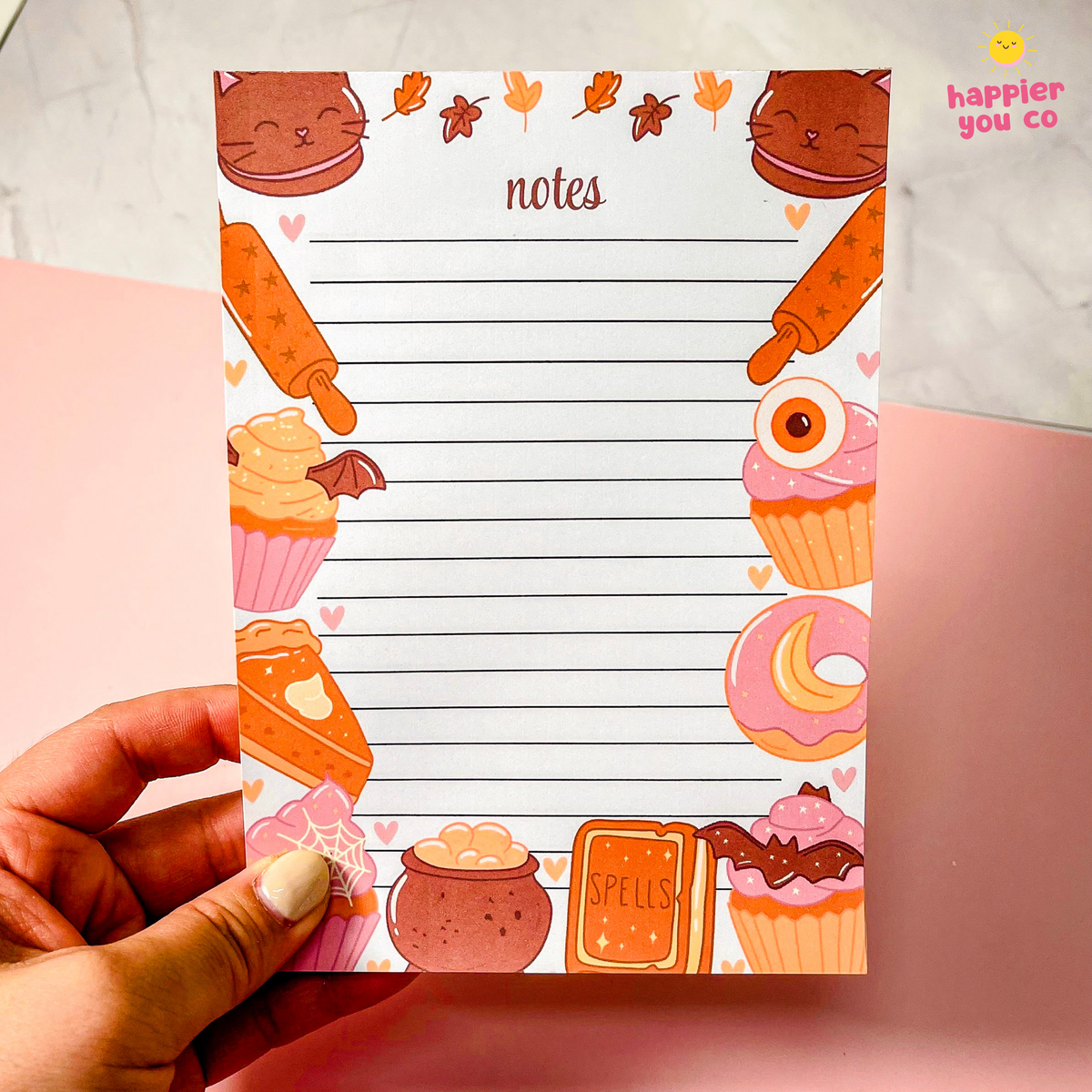A hand holds a cute autumn-themed notepad with lined writing space and illustrations of cupcakes, cookies, leaves, and a cauldron. The notepad has a white background with orange, pink, and brown seasonal doodles.