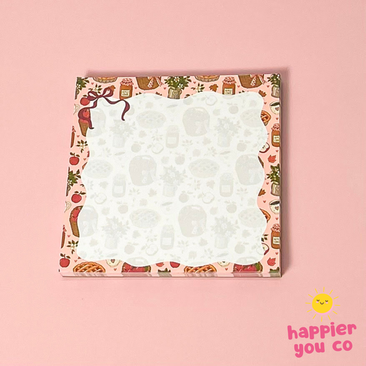 A square notepad with a pink kitchen-themed border and a white center, photographed on a solid pink background. A small 'happier you co' logo appears in the bottom right corner.
