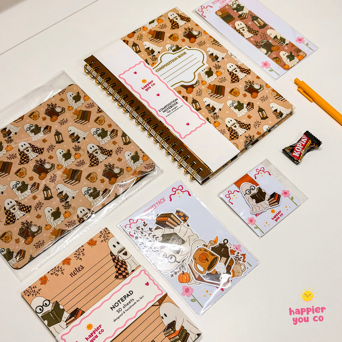A Halloween-themed stationery set arranged on a white surface, including a spiral notebook, memo pads, a sticker sheet, and a small candy wrapper. The stationery features a warm autumn palette with ghosts, pumpkins, leaves, and candy motifs.