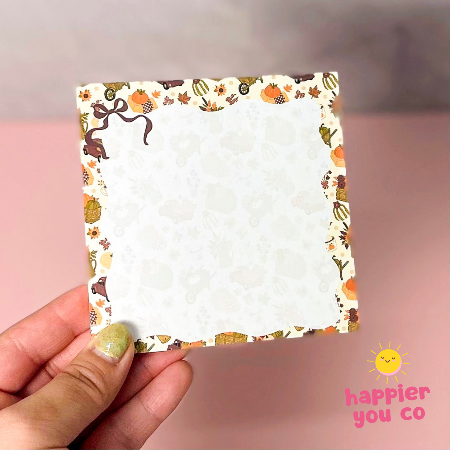 A hand holding a square white notepad with a decorative autumn-themed border featuring pumpkins, leaves, and bows, against a soft pink background with a 'happier you co' logo in the bottom right corner.