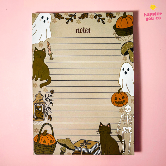 A Halloween-themed lined notes pad with cute illustrations of ghosts, pumpkins, a black cat, a skeleton, a lantern, and mushrooms around the border, photographed on a pink background. The brand name 'happier you co' appears in the top right corner.