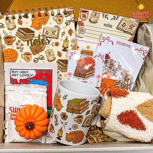 A cozy autumn-themed stationery and mug gift set arranged in a box. The set includes a spiral-bound notebook, a sticky notes pad, a sticker sheet, a white ceramic mug with fall illustrations, a small orange pumpkin decoration, a Swig Puf tea bag packet, and a cream knit blanket with orange and brown patterns.