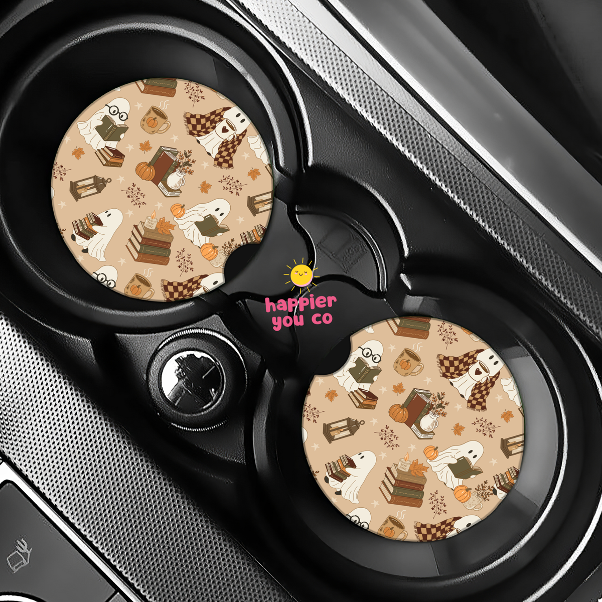 Two round car cup holder inserts with a beige Halloween pattern featuring ghosts, pumpkins, and treats, installed in a black car center console. The center hub has the pink text 'happier you co' with a small yellow sun graphic.