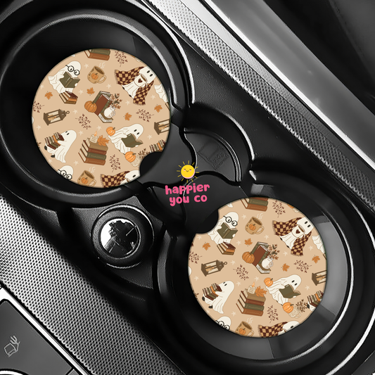 Two round car cup holder inserts with a beige Halloween pattern featuring ghosts, pumpkins, and treats, installed in a black car center console. The center hub has the pink text 'happier you co' with a small yellow sun graphic.