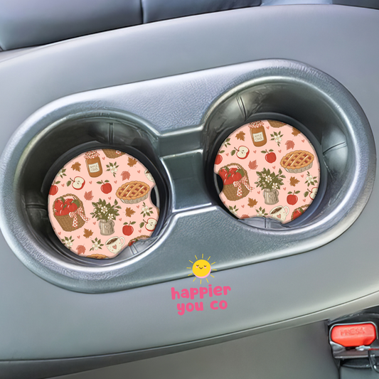 Two circular car cup holders on a gray vehicle center console, each holding a pink fall-themed coaster with a pumpkin, acorn, apple, pie, and leaves pattern. A small 'happier you co' logo appears near the bottom center.