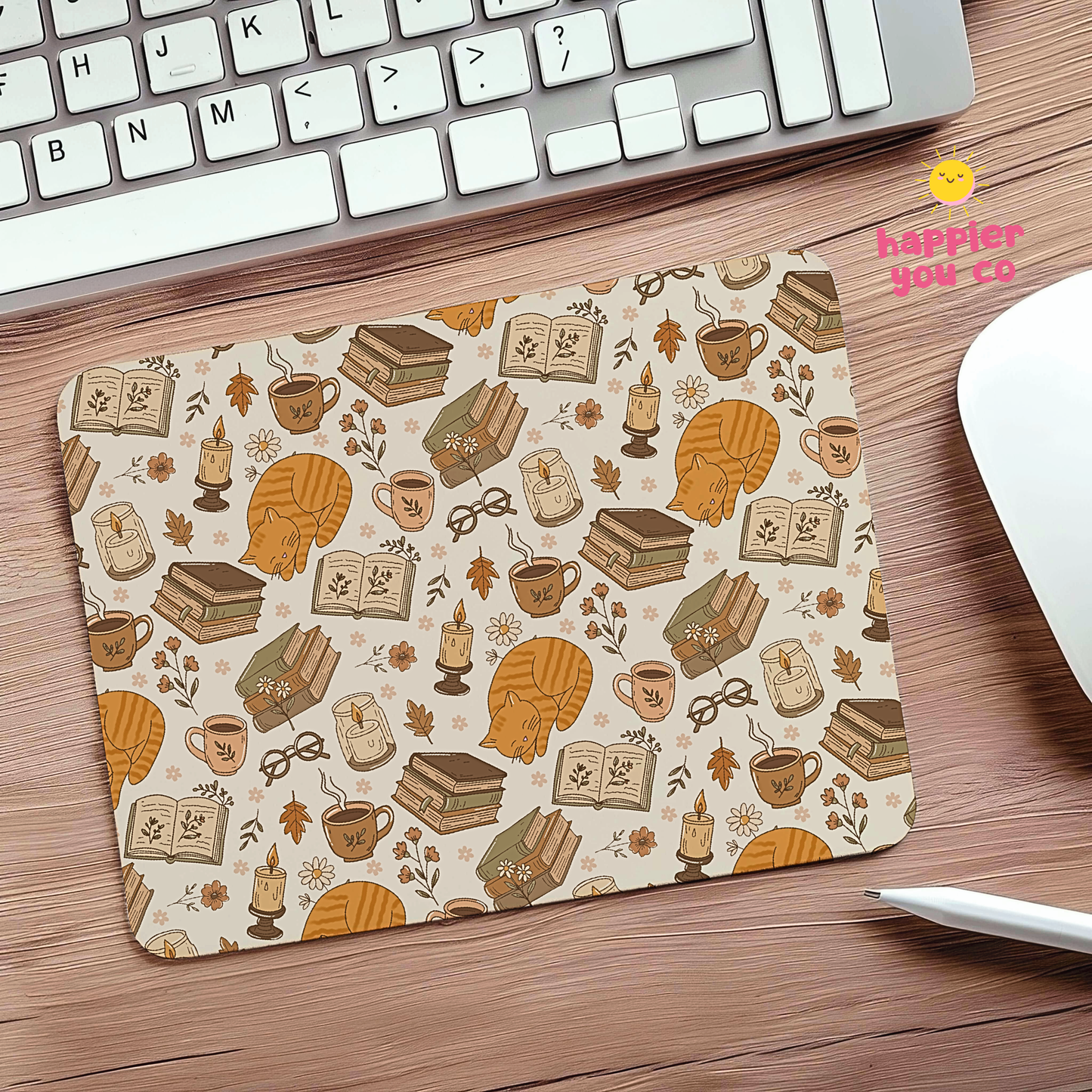 A rectangular desk mat with a cozy autumn-themed pattern of books, mugs, candles, leaves, and pumpkins on a cream background, placed on a wooden desk next to a keyboard and mouse.