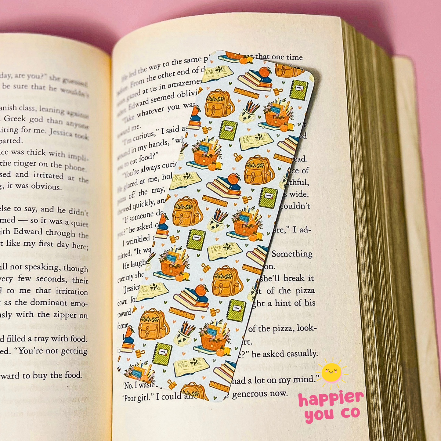 A long, narrow bookmark or sticker with a colorful cartoon school-themed pattern is placed on an open book page. The pattern shows a smiling orange cat, backpack, books, pencils, and school supplies.