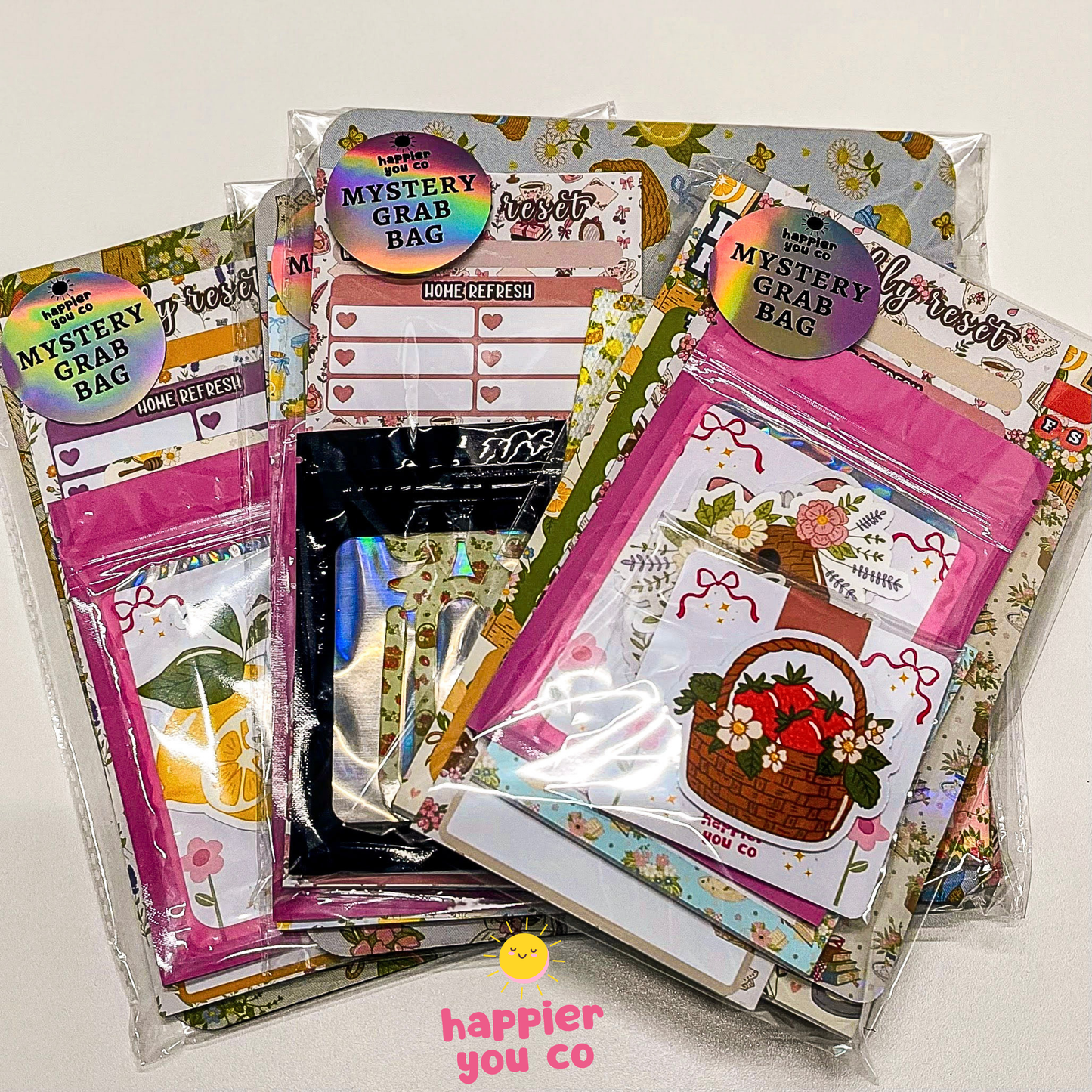 Three mystery grab bags of stationery and craft supplies displayed together on a light surface. The bags feature colorful floral and patterned designs with stickers, pens, and decorative items visible inside.