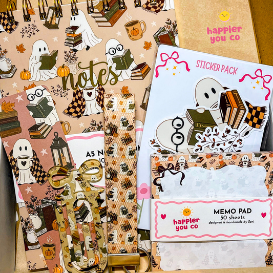 A styled flat-lay of a stationery gift set from Happier You Co, featuring a ghost and book patterned notebook, a sticker pack, a memo pad, and a patterned pen loop inside a kraft paper box.