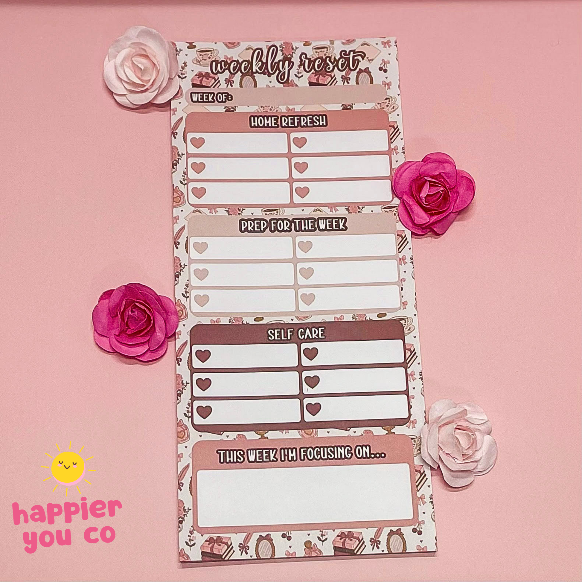 Weekly planner notepad with floral design on a pink background, featuring the 'happier you co' brand.
