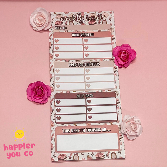 Weekly planner notepad with floral design on a pink background, featuring the 'happier you co' brand.